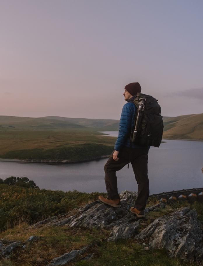 Man with a backpack on his back, stood on a mountain edge looking out across a landscape of mountains and a lake.