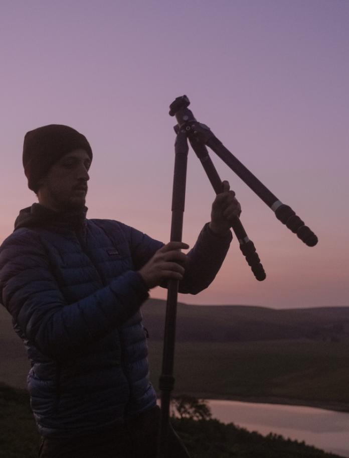 Man holding up a camera tripod silhouetted against a pink and purple sunset sky, 