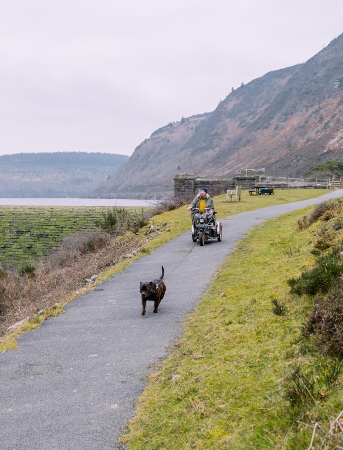 Eine Frau mit Elektromobil zusammen mit Hund auf einem geteerten Weg an einem Stausee.