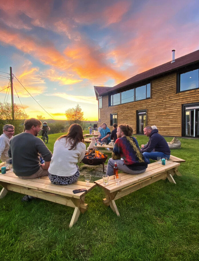 A group round a firepit outside a chalet-style building at sunset.