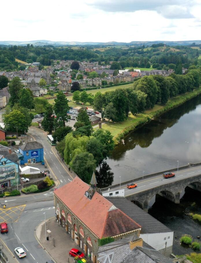 A birds eye view of Builth Wells. The old stone bridge crosses a river lined with green trees. Roofs of the town buildings can be seen.