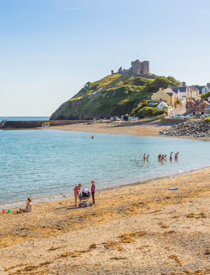 Criccieth seafront with golden sand and people enjoying themselves on the beach. The sea and sky are blue and Criccieth Castle can be seen on a rock in the distance.