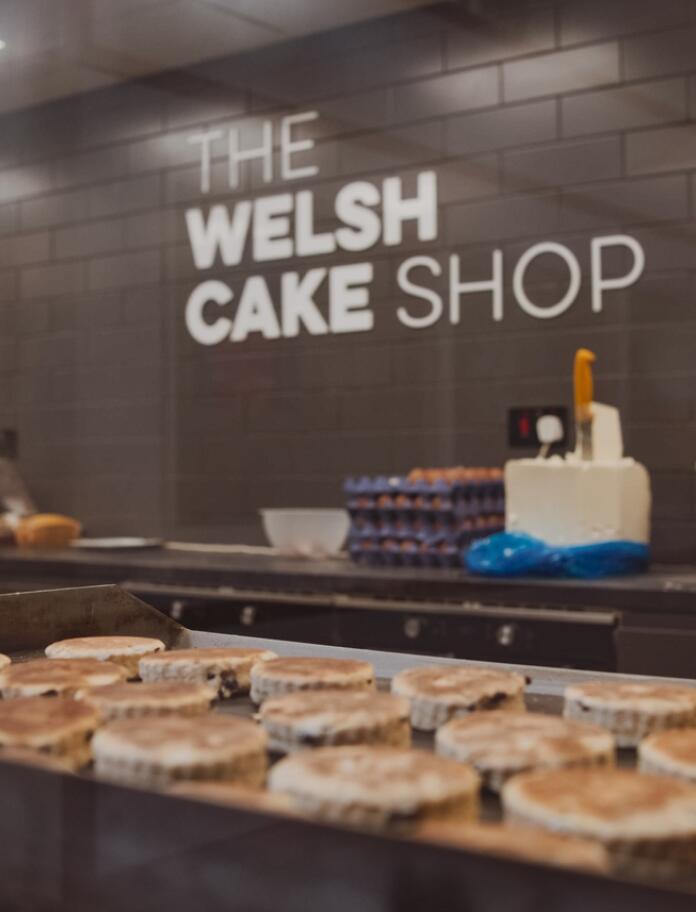 A welsh cake stall at a market with The Welsh Cake Shop sign in the background. Welsh cakes are being cooked on the griddle in the foreground. 