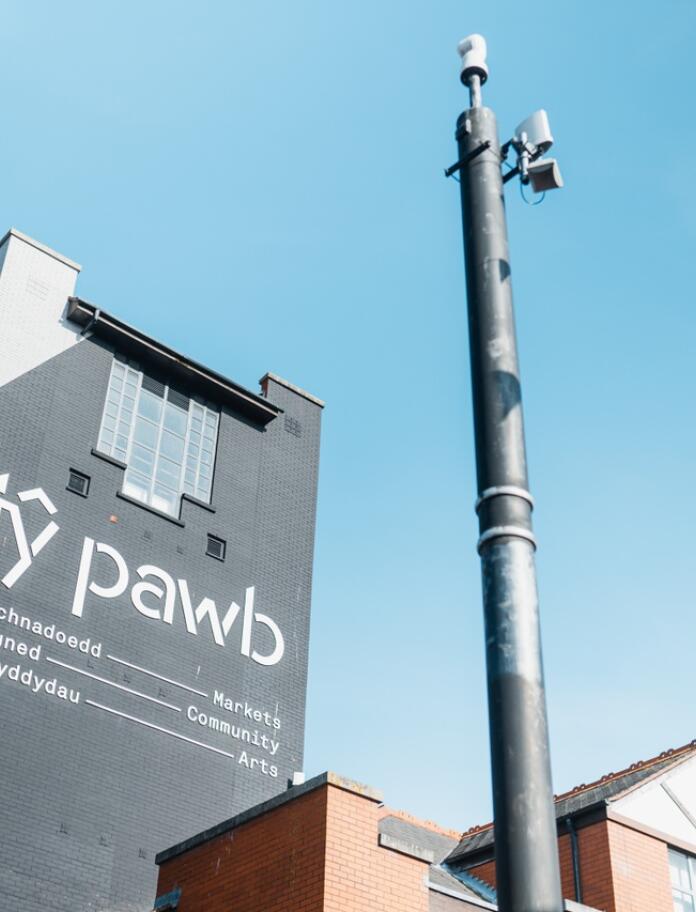 Exterior shot of Tŷ Pawb art centre - the sign on the tall grey building says 'market, community, arts'. The sky is blue and there's bunting swaying in the wind next to the building. 