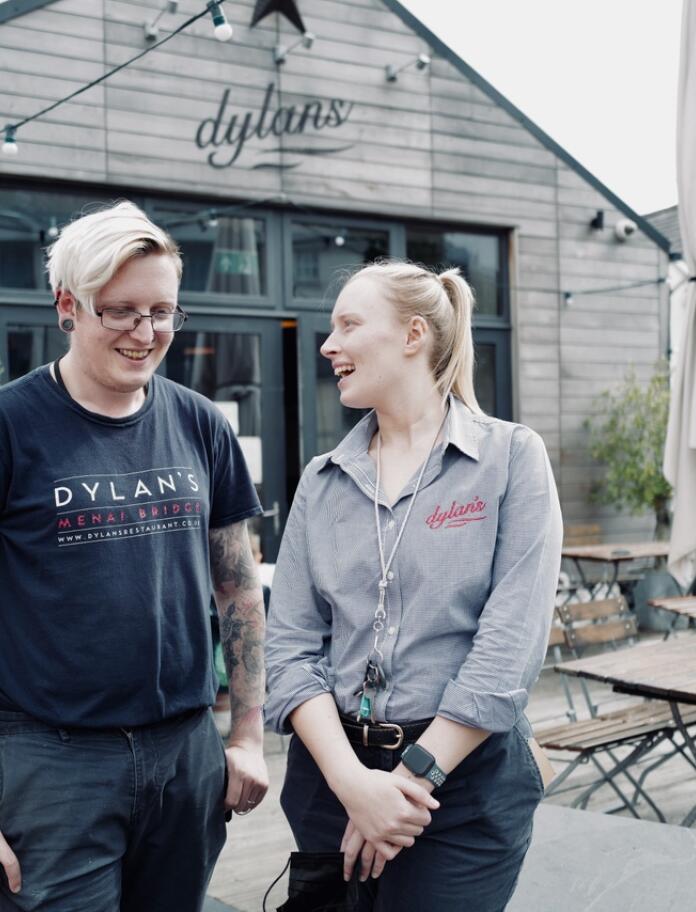 Two people standing outside a wooden restaurant smiling. Both work at the restaurant and are wearing an uniform with 'Dylan's Restaurant' printed on.