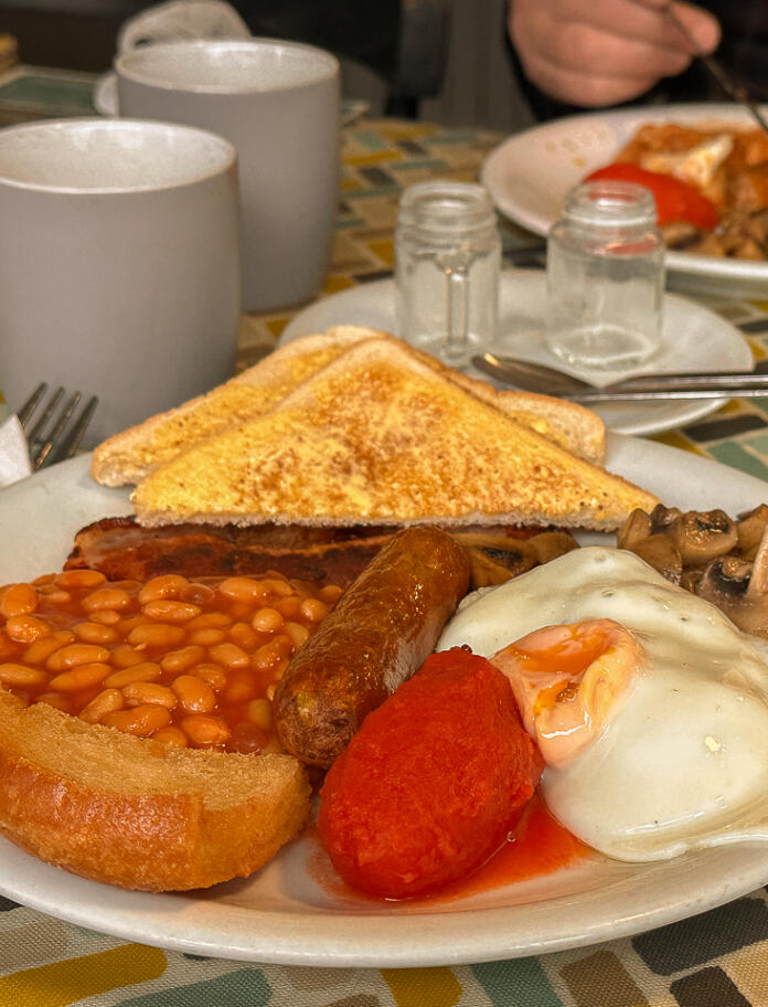 Ein Frühstücksteller mit Würstchen, gebackenen Bohnen, Tomaten, Speck, Spiegelei, Champignons, Toast und einer Tasse Tee.