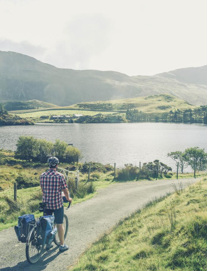 Ein Radfahrer in kariertem Hemd der auf einem Weg steht und auf einen See mit Bergen im Hintergrund blickt.