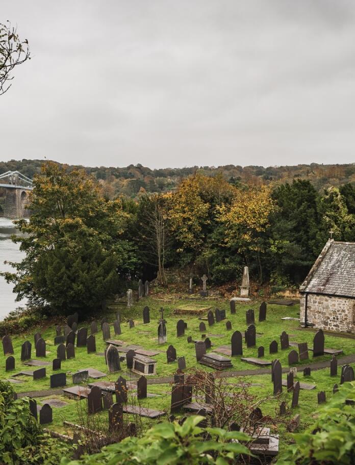 A small chapel with a waterfront cemetery and a bridge in the background.