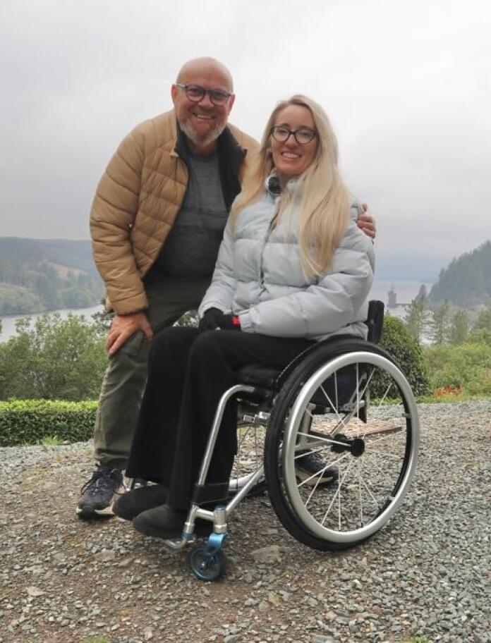 A standing man and a woman in a wheelchair with a large lake in the background.