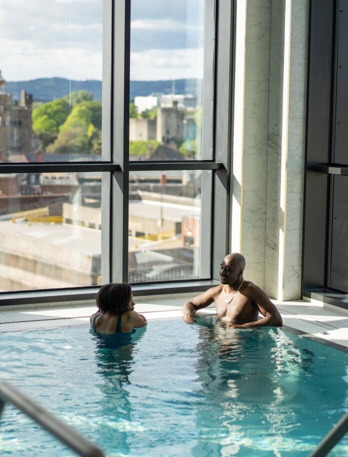 Two people relaxing in a spa pool with views through a large window over a city.