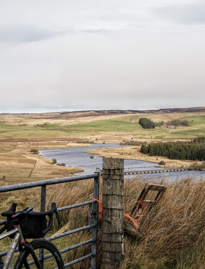 A bike resting on a metal farm gate, overlooking a lake in hills.