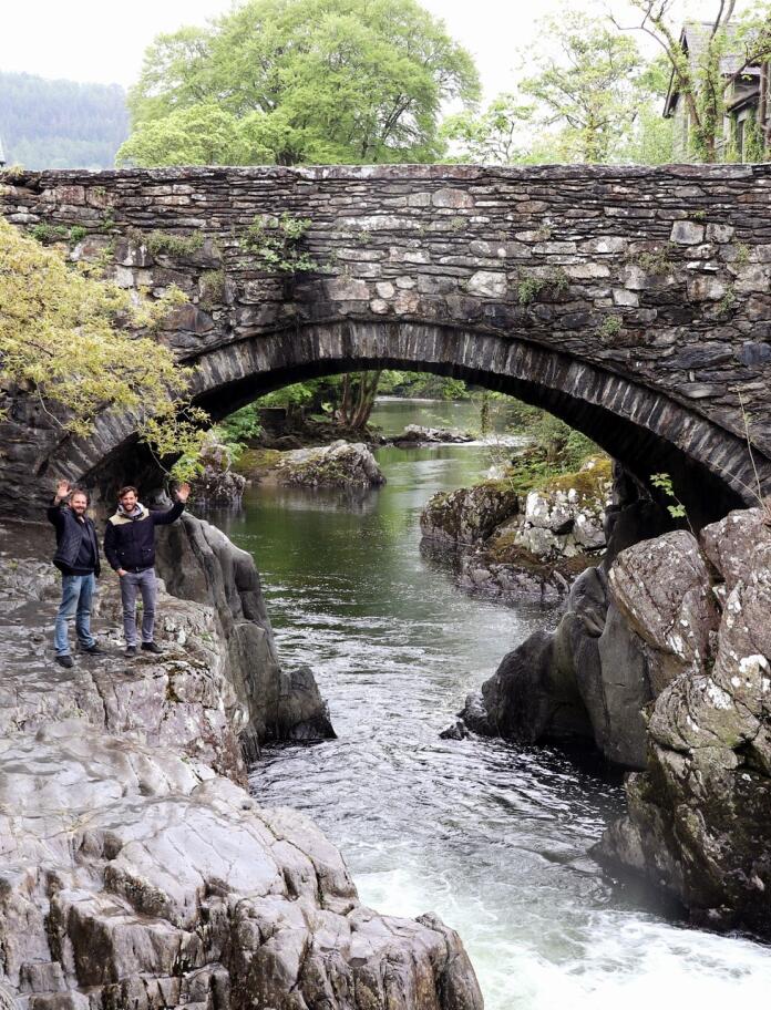 A grey stone bridge over a narrow river and two men waving.