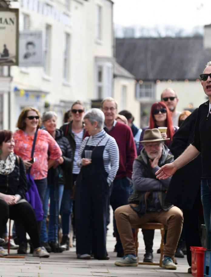 Eine Gruppe Personen, sitzend und stehend, die einem Vortrag auf der Straße lauschen.