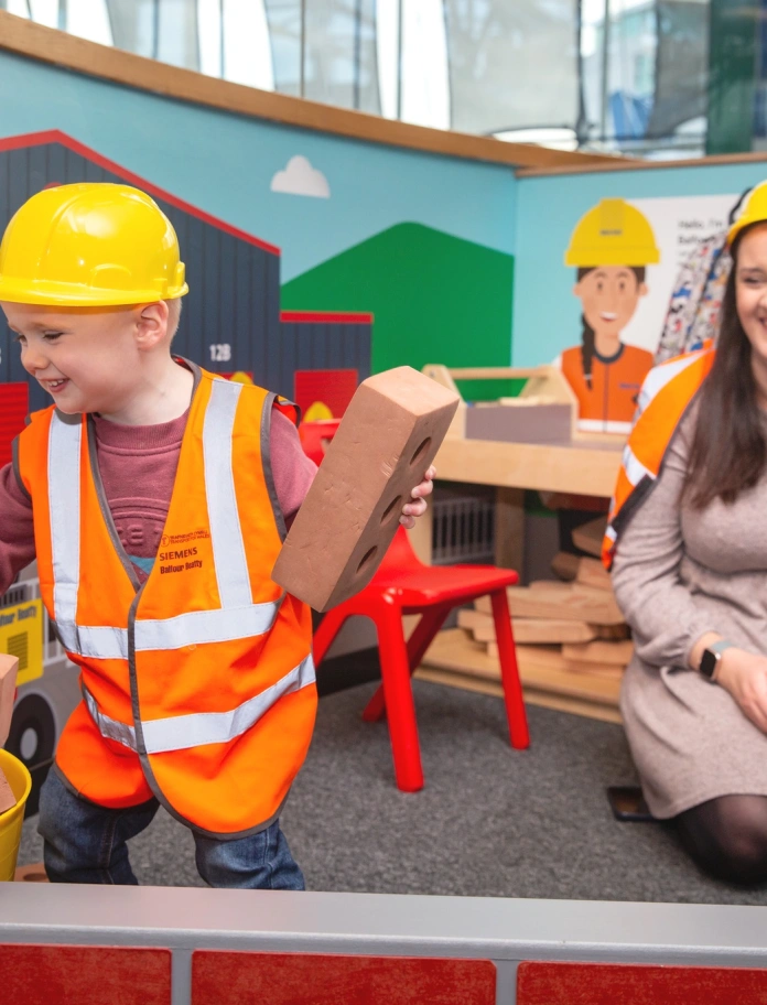 boy with helmet and hi vis jacket playing with bricks as woman watches.