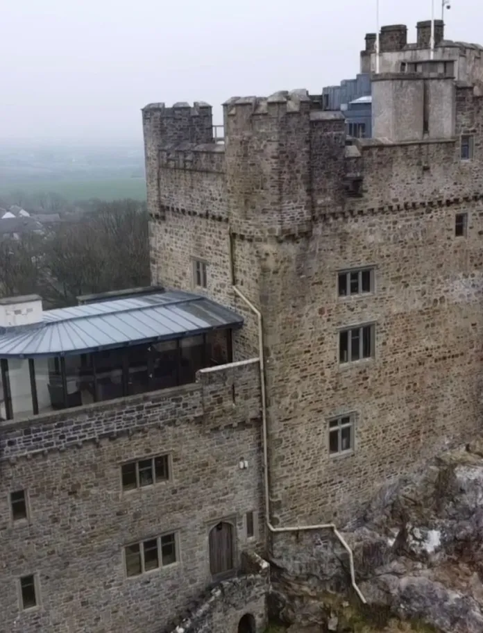 Aerial view of a historic stone castle with a modern glass extension, surrounded by a rural landscape and several nearby buildings.