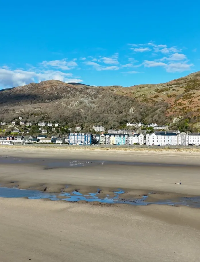 A seaside town with a wide sandy beach, colourful houses along the seafront and a backdrop of green hills behind.