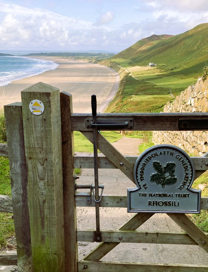 Hölzernes Tor mit Schild Rhossili und einem langen Sandstrand im Hintergrund.