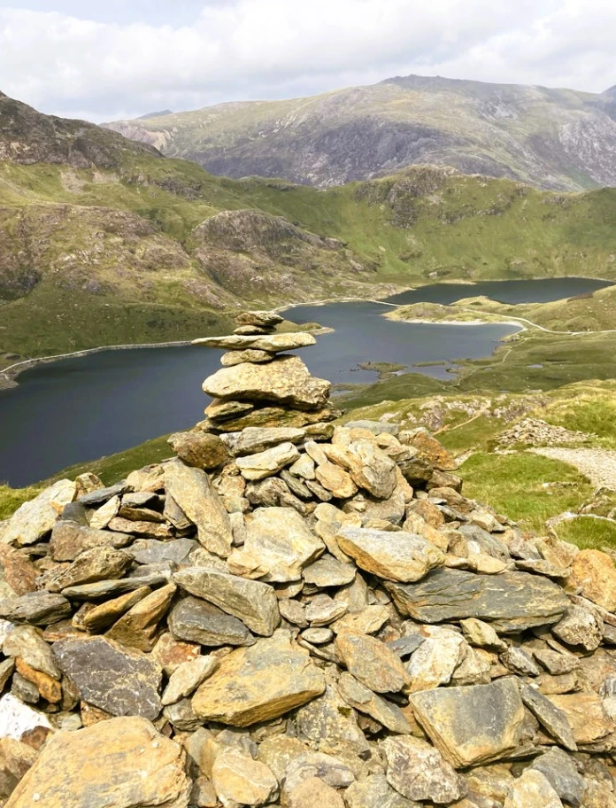 Ein Bergsee mit Steinhaufen im Vordergrund und hohen Gipfeln.