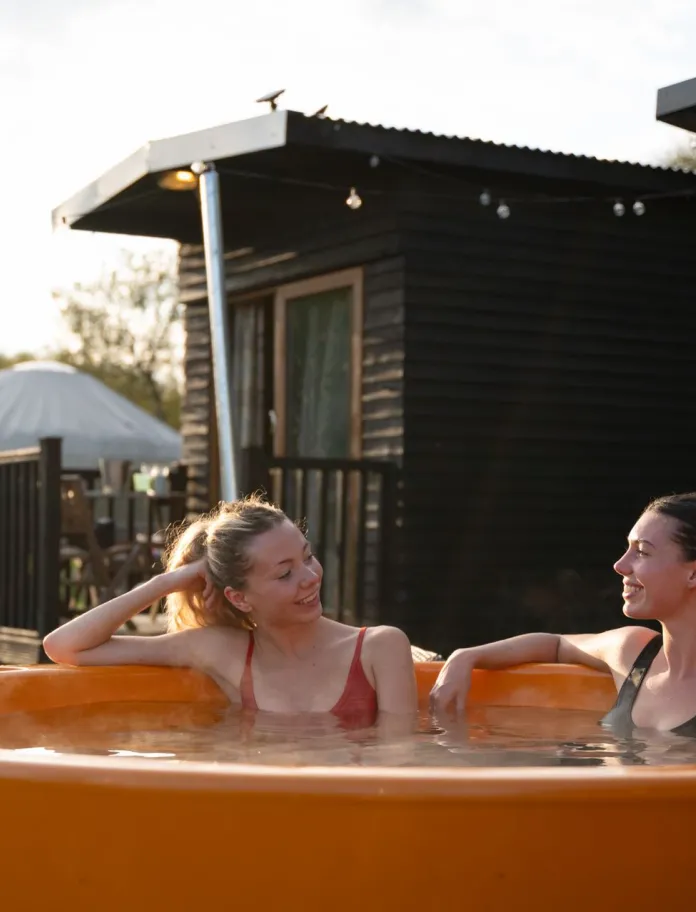 Two people sitting in a round, bright orange outdoor hot tub at a rustic retreat. Behind them is a small dark wooden cabin with a pitched roof and a white tent structure in the background. The setting includes grassy terrain and natural greenery under soft daylight, creating a relaxed outdoor spa atmosphere.