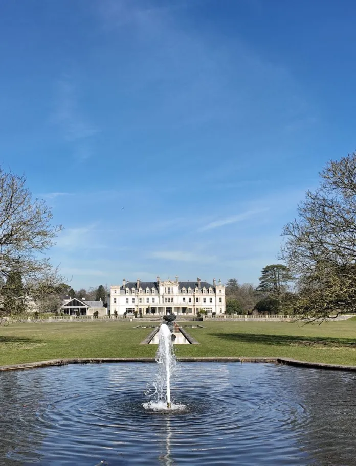 A mansion house set in large gardens, with a fountain in the foreground.