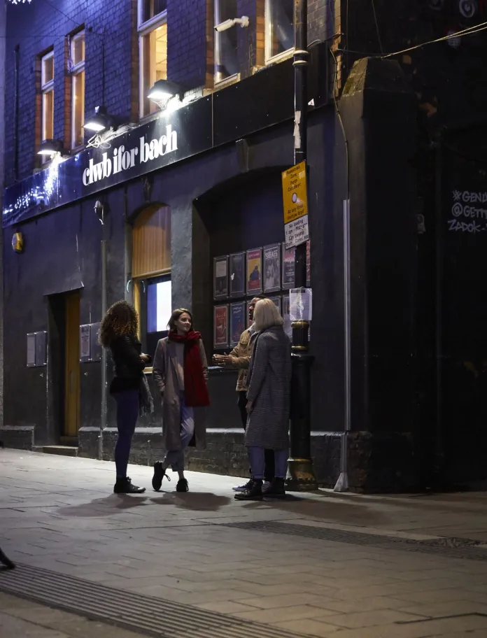 A group of people standing in a street outside a music club at night.