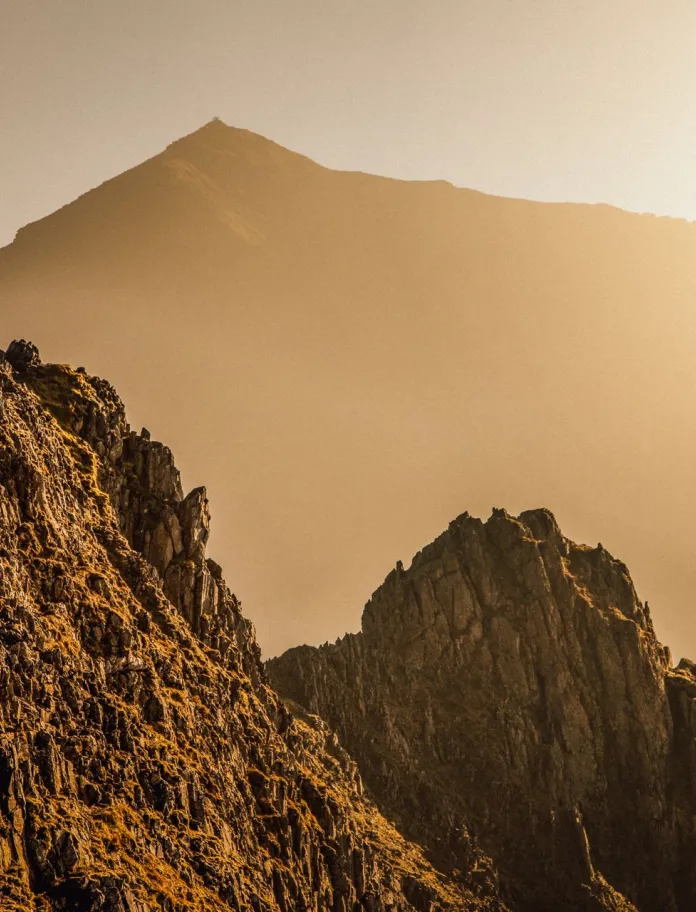 A sunrise view over a rugged mountain landscape, with lakes and ridges partly covered by low cloud and a small lone figure standing on a rocky foreground slope.