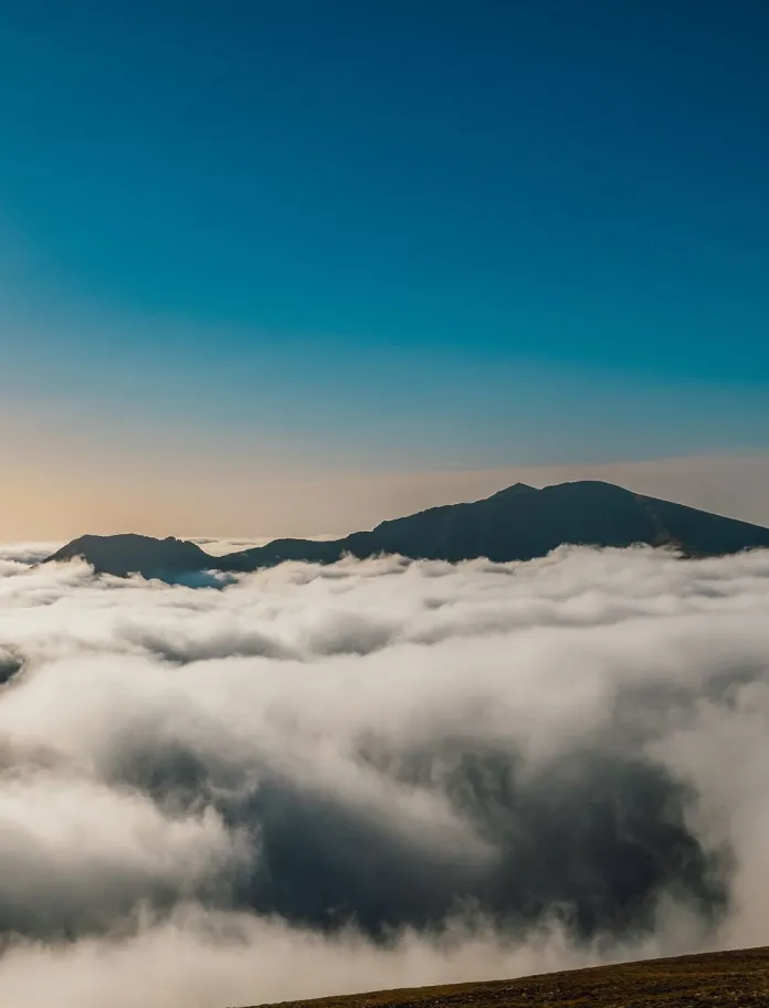 A lone walker stands on a ridge above a thick cloud inversion, with distant mountain peaks rising through the clouds under a clear blue sky.