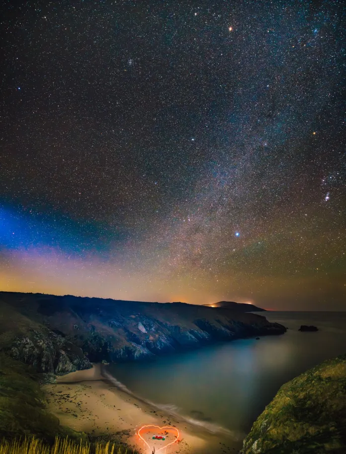 A quiet beach enclosed by cliffs at night, with a small circle of warm light on the sand beneath a wide sky filled with stars and the Milky Way.
