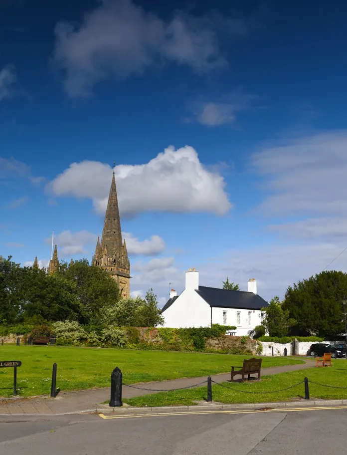 Cathedral green in Llandaff with a tall spired cathedral behind trees, a statue on a plinth to the left, a white house to the right, and a cyclist passing on the road under a blue sky.