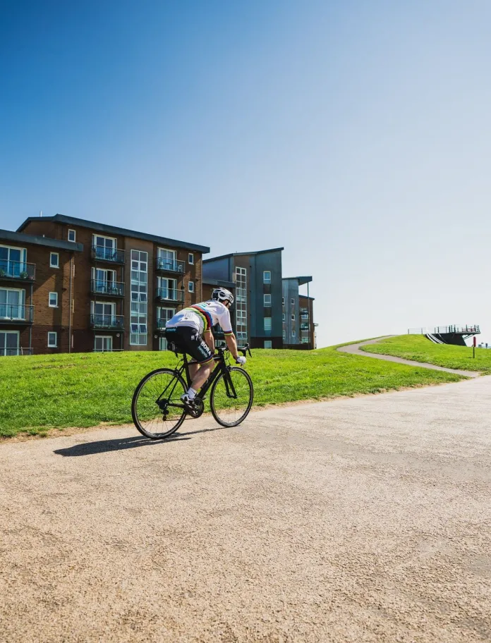 Ein Radfahrer auf einem Radweg mit Gebäuden und Rasen davor und blauem Himmel.