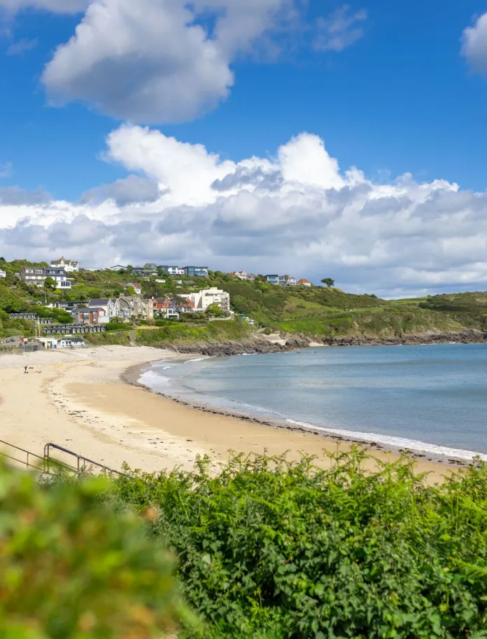 A sandy bay with calm sea, hillside houses, and a bright blue sky with clouds.