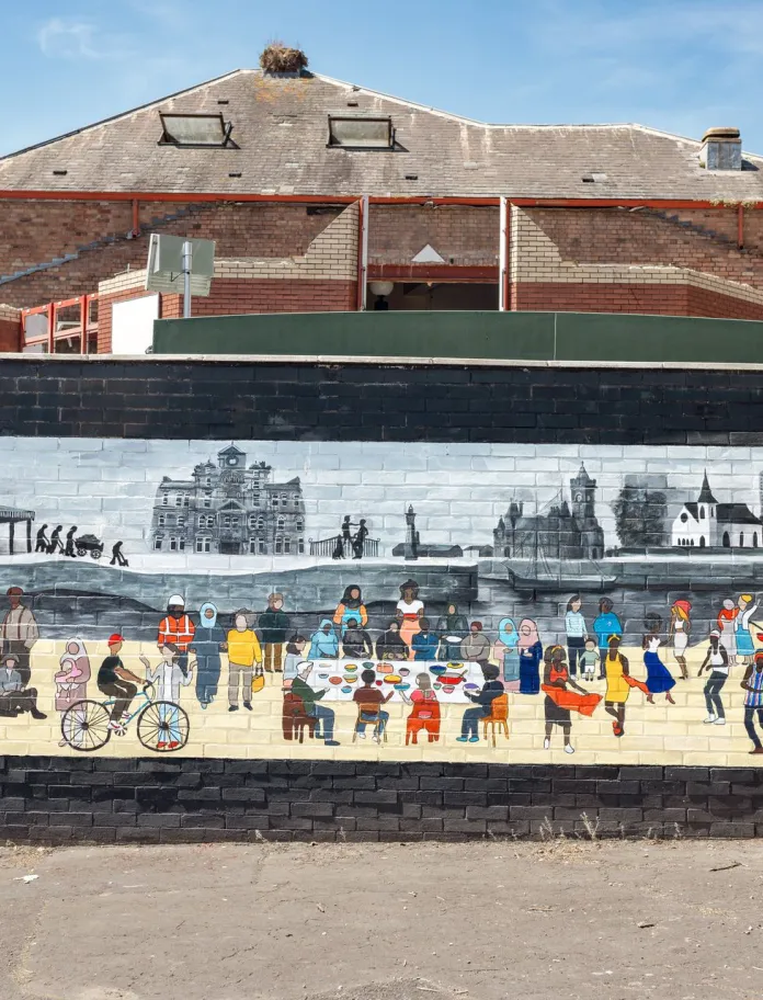 Community mural depicting people gathering, eating, cycling and taking part in activities, set against a monochrome Cardiff Bay skyline painted on a brick wall.