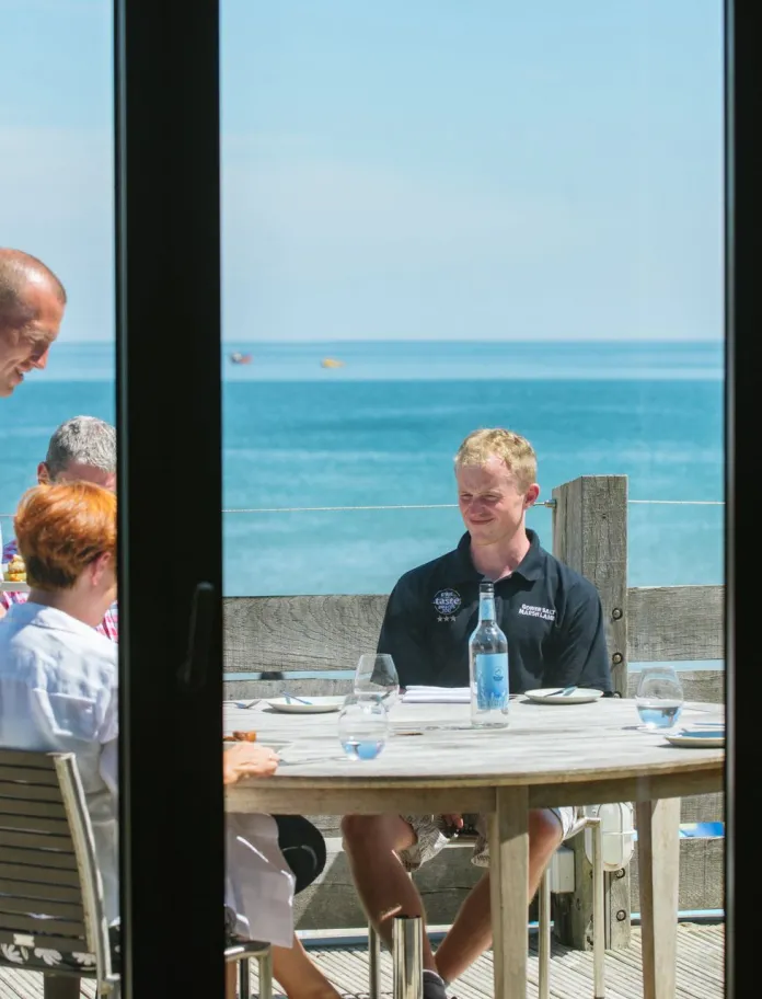 View through a window towards an outdoor seaside terrace, with people standing and sitting around wooden tables overlooking the coast on a bright day.
