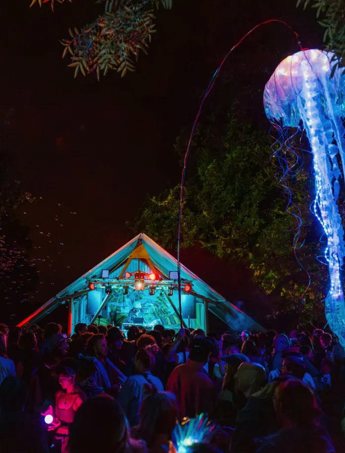 A festival at night, with glowing jellyfish floating in the sky and people partying in front of a tent.