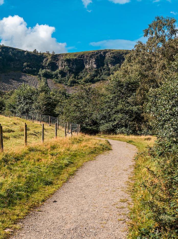 Ein Wanderweg rund um ein Feld mit einem grünen Steilhang im Hintergrund.