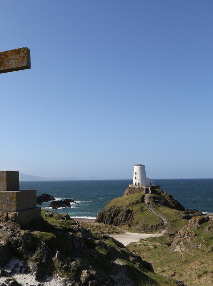 Steinkreuz mit einem Leuchtturm und dem Meer im Hintergrund.