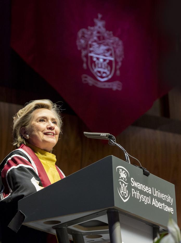 woman wearing gown speaking at podium with university logo and name  with university logo projected onto wall behind
