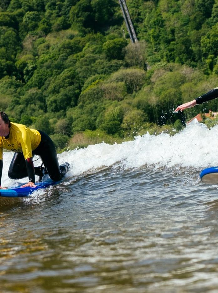 Two people wearing black wetsuit with a yellow t shirt on surfboards in the water
