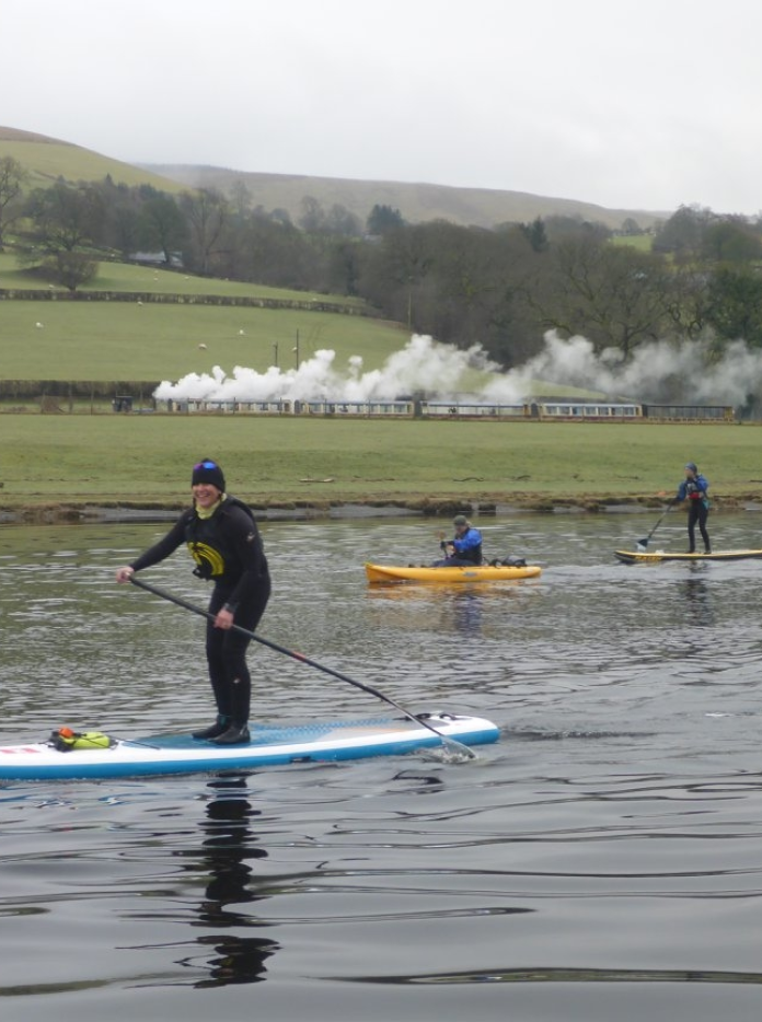 Menschen auf Stand Up Paddle Boards mit Dampfzug im Hintergrund.