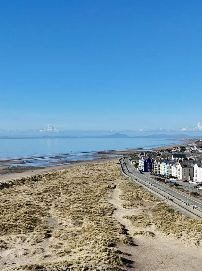 Sandy beach and promenade at Harlech with dunes, a long walkway, and mountains in the background under a clear blue sky.