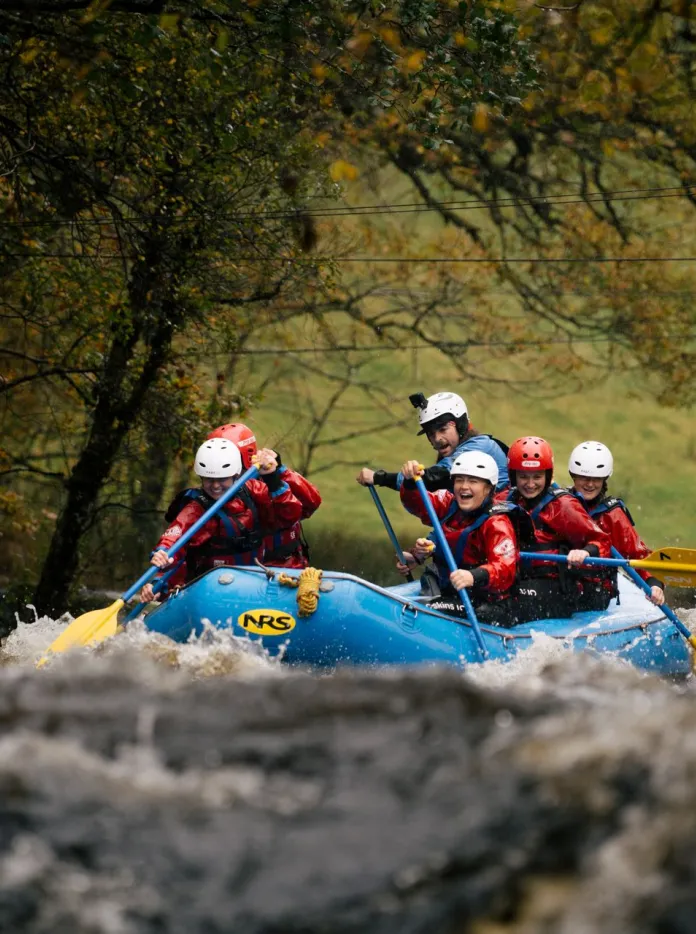 People white-water rafting in a blue inflatable raft on a fast-flowing river, wearing safety helmets and buoyancy aids, with trees and countryside in the background.
