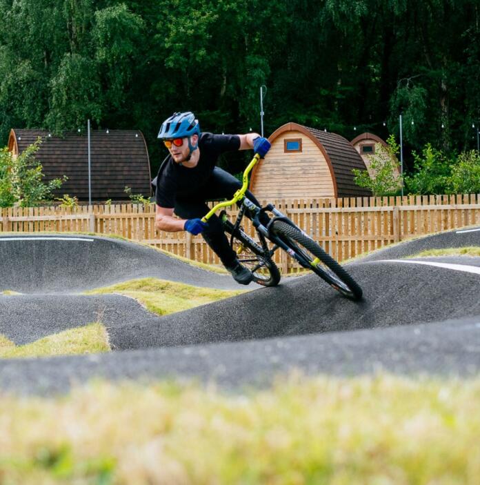 A mountain biker cycling around a pump track with glamping pods in the background.