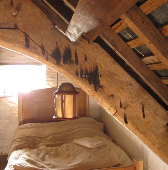 Ein altes Dachgeschosszimmer mit einem kleinen Fenster, Balken und einer Laterne über dem Bett.