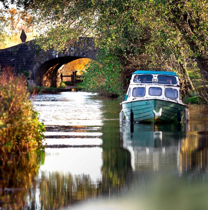 Boat on the canal moored under a tree with a bridge in the background.