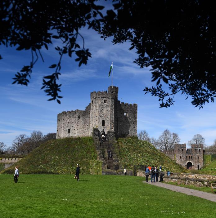 People walking up to The Keep, a tower at Cardiff Castle framed by a tree in the foreground.
