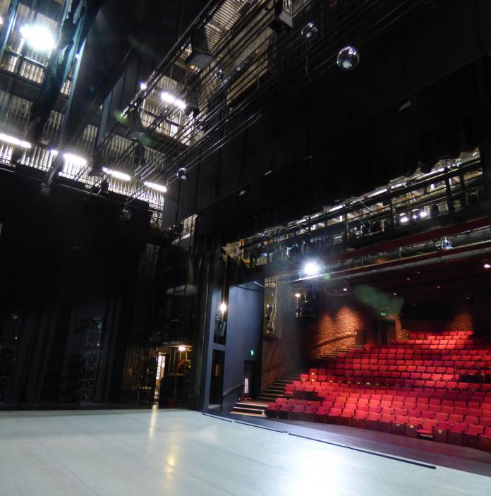 Auditorium mit roten Sitzen und Bühne, von den Seitenflügeln aus gesehen.