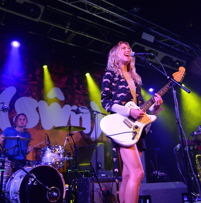 A woman playing guitar and singing on stage with a drummer in the background.