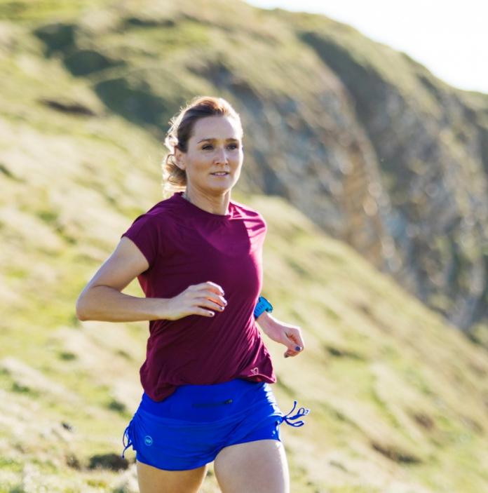 A woman wearing a red top and blue shorts running across a grassy cliff
