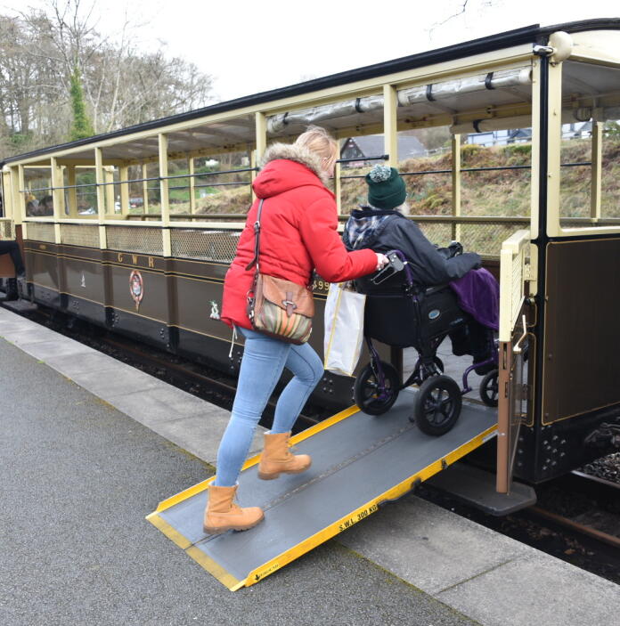 A lady pushing a person in a wheelchair up a ramp into a narrow gauge train carriage.