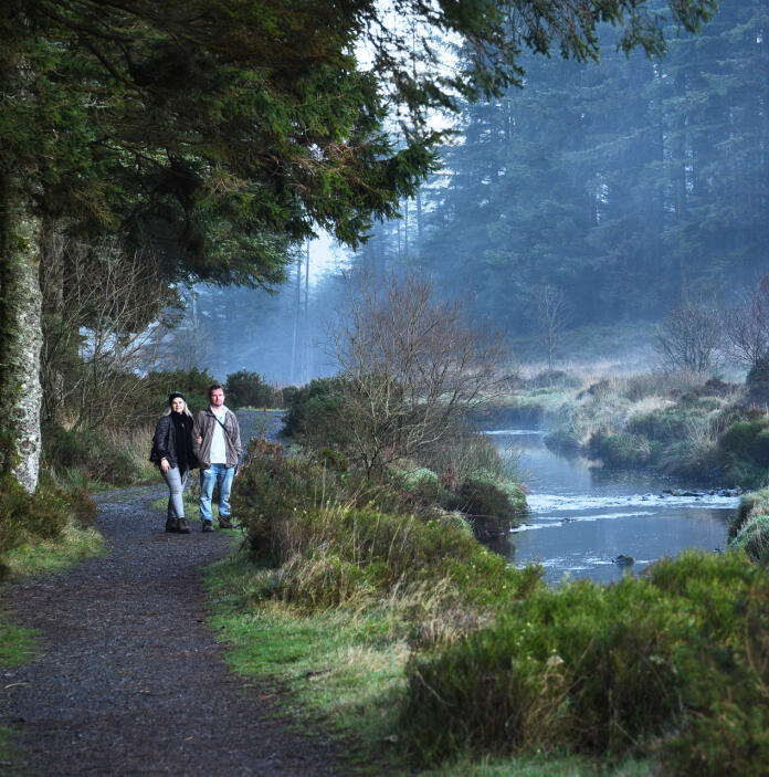 Two people walking in a forest, alongside a river. 